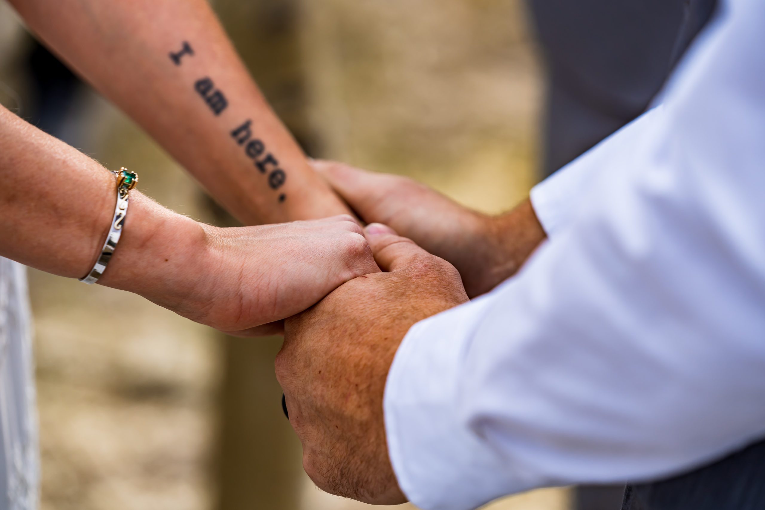 A bride with a tattoo on her arm holds hands with her groom during their backyard wedding ceremony