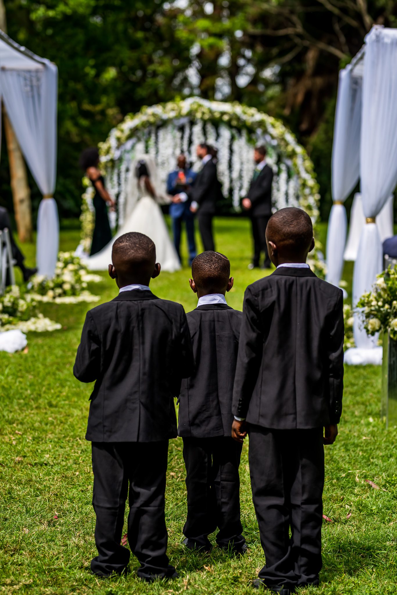 three young ring bearers stand facing away from the camera and looking down the isle at a wedding