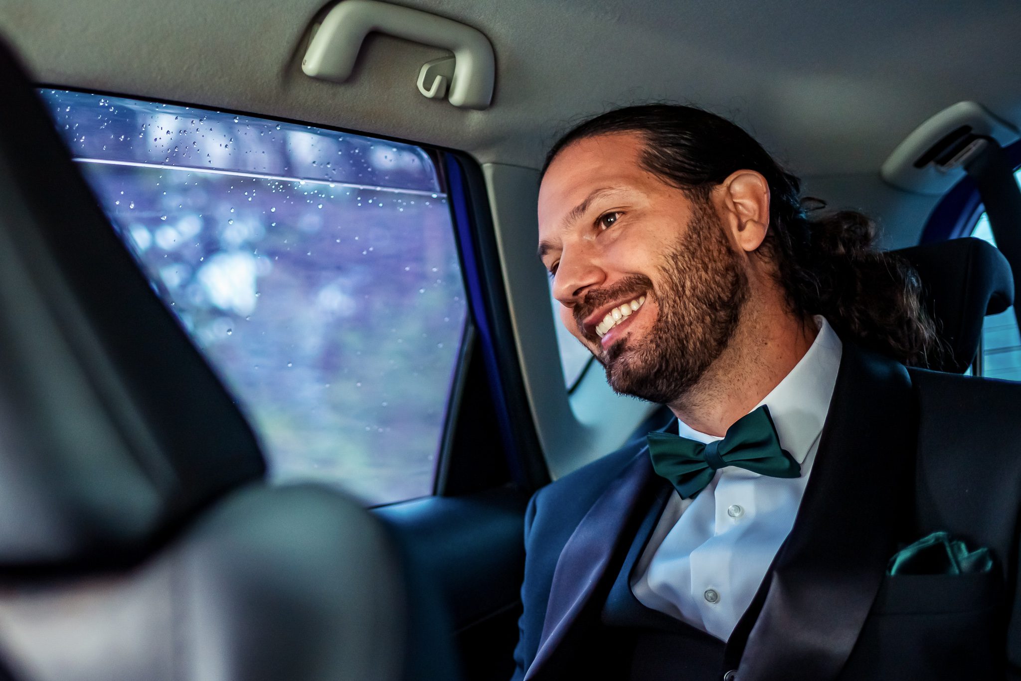 a groom smiles as he sits in the back of a car on the way to his wedding