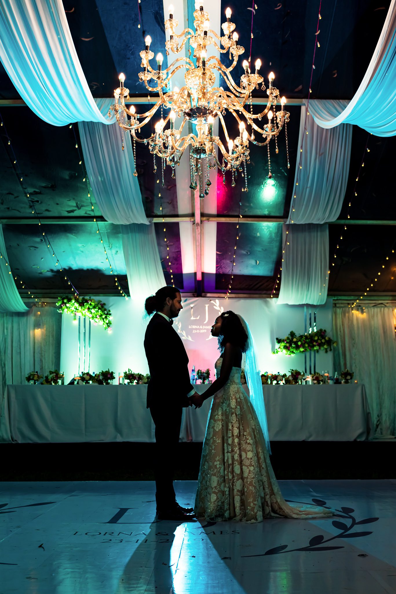 two figures in silhouette hold hands and face each other. one is in a wedding dress and the other in a tux. they stand below a beautiful chandelier in a room with multicolored lights illuminating the wedding venue background