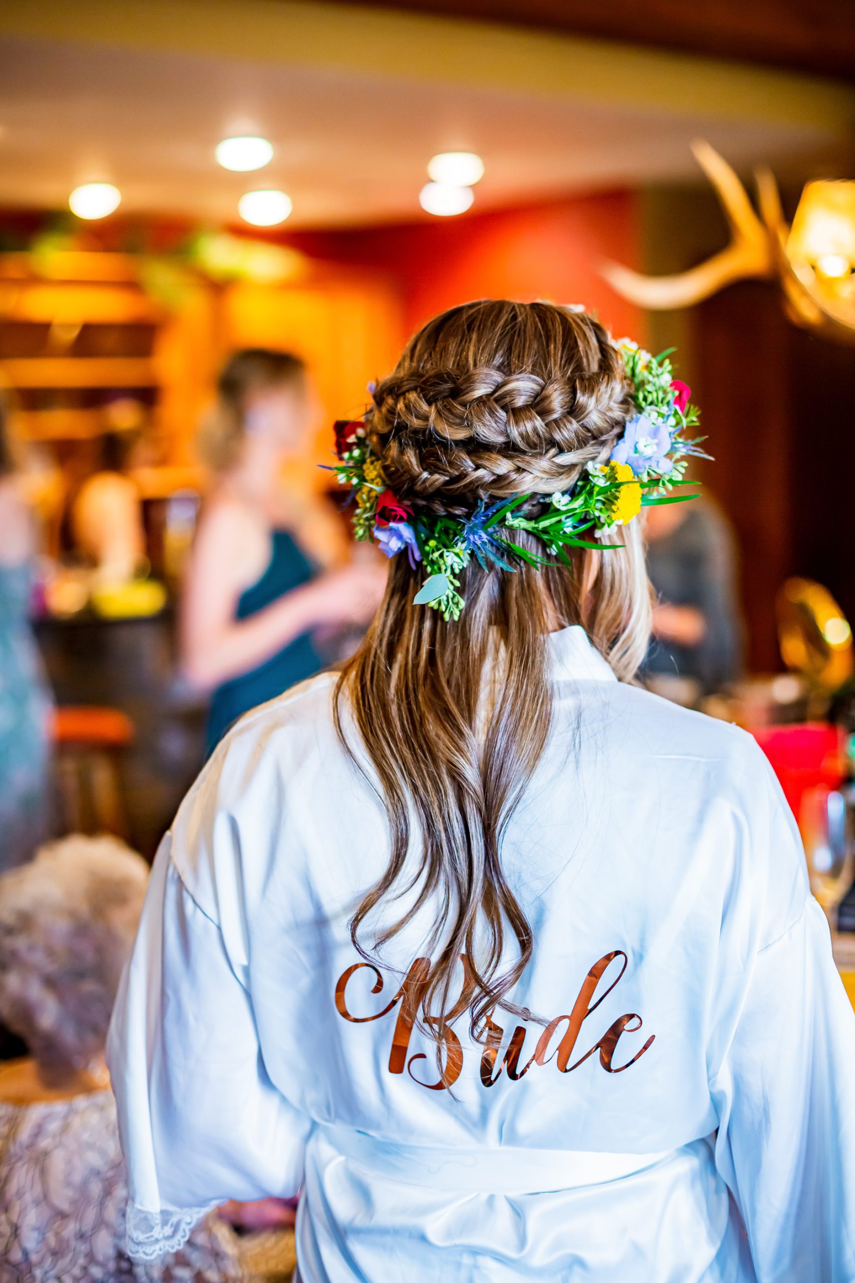 A blonde bride wearing a flower crown and a white robe that says bride on the back.