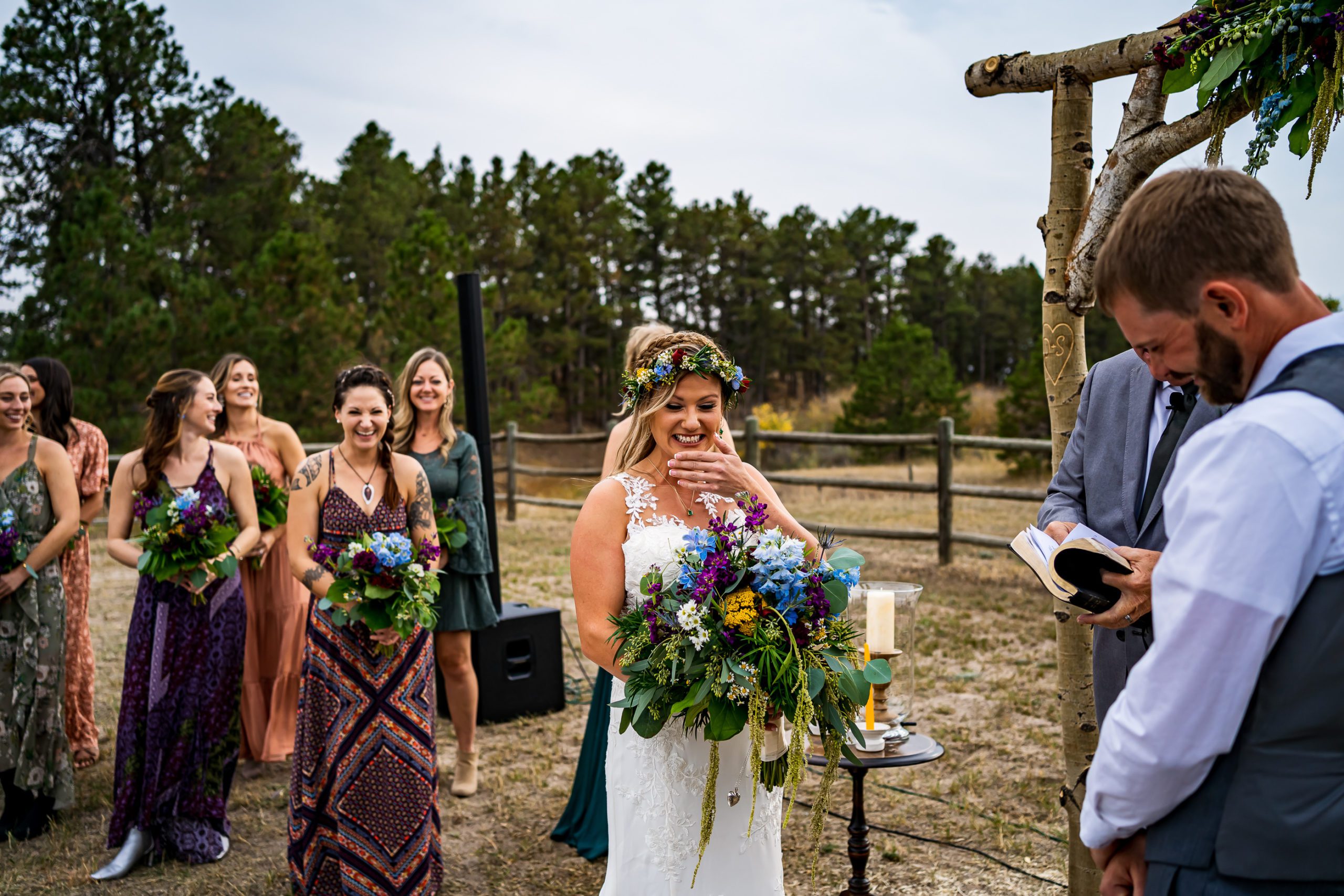 A bride stands in front of her bridesmaids holding a large bouquet of blue and yellow flowers at her Colorado backyard wedding.